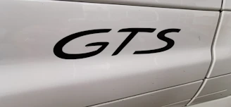 The image features a close-up view of the word 'GTS' in black lettering on the side panel of a white vehicle. The background shows smooth, glossy paint with light reflections and some visible panel lines.