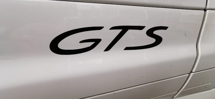 The image features a close-up view of the word 'GTS' in black lettering on the side panel of a white vehicle. The background shows smooth, glossy paint with light reflections and some visible panel lines.