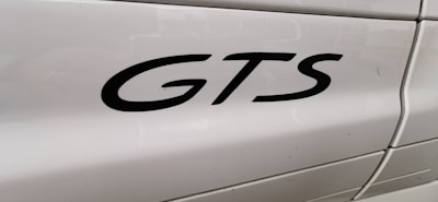 The image features a close-up view of the word 'GTS' in black lettering on the side panel of a white vehicle. The background shows smooth, glossy paint with light reflections and some visible panel lines.