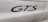 The image features a close-up view of the word 'GTS' in black lettering on the side panel of a white vehicle. The background shows smooth, glossy paint with light reflections and some visible panel lines.