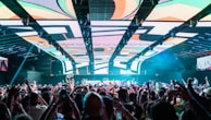 A lively crowd packed under a ceiling with bright, abstract designs, suggests an energetic concert or club scene. People are enthusiastically raising their hands and recording with their phones, with colorful lighting enhancing the vibrant atmosphere.