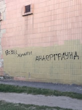 Graffiti is sprayed in black paint on a tiled wall. The wall consists of bricks in beige and light peach colors. There is a vent visible, and the base of the wall meets a patch of grass and a dirt path.