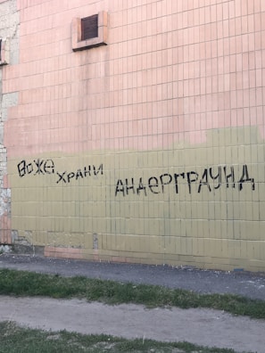 Graffiti is sprayed in black paint on a tiled wall. The wall consists of bricks in beige and light peach colors. There is a vent visible, and the base of the wall meets a patch of grass and a dirt path.