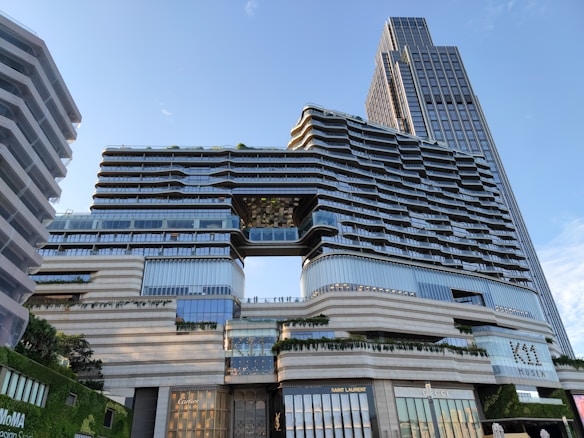 A modern multi-story architectural building features a distinctive, wavy facade with glass and metallic elements. The structure includes a suspended section connecting two parts of the building. Prominent luxury brand stores like Cartier, Saint Laurent, and Gucci are located on the ground floor. The setting includes greenery integrated into the design, and a clear sky in the background.