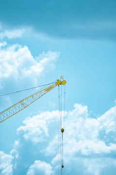 A yellow construction crane stands prominently against a backdrop of bright blue sky filled with fluffy white clouds. The crane's arm extends diagonally across the image, with cables hanging downward.