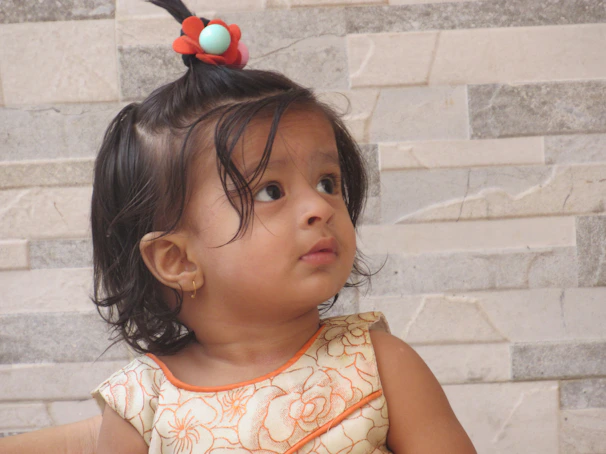 A young child with dark hair tied up with a red and blue hair accessory, wearing a floral-patterned dress with orange accents, is looking to the side. The background consists of a light-colored tile wall with a subtle pattern.