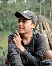 A young person wearing a dark cap and sweatshirt is sitting outdoors in a natural setting. They appear thoughtful, with their hand raised to their chin and a slight smile on their face. The background consists of blurred greenery and what appears to be wooden logs or rocks.