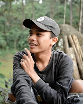 A young person wearing a dark cap and sweatshirt is sitting outdoors in a natural setting. They appear thoughtful, with their hand raised to their chin and a slight smile on their face. The background consists of blurred greenery and what appears to be wooden logs or rocks.