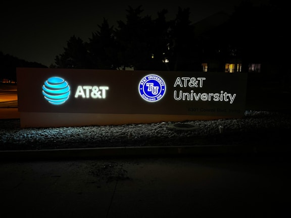 An illuminated signboard displays the AT&T logo, the wording 'AT&T University,' and an emblem with the text 'AT&T University Established 2009.' The sign is positioned outside against a background of trees, and the setting appears to be at night.