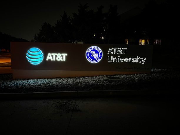 An illuminated signboard displays the AT&T logo, the wording 'AT&T University,' and an emblem with the text 'AT&T University Established 2009.' The sign is positioned outside against a background of trees, and the setting appears to be at night.