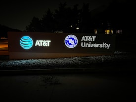 An illuminated signboard displays the AT&T logo, the wording 'AT&T University,' and an emblem with the text 'AT&T University Established 2009.' The sign is positioned outside against a background of trees, and the setting appears to be at night.