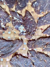 Slices of dried meat arranged on a woven bamboo mat, sprinkled with chili flakes and seasonings. The texture of the meat is rugged and fibrous.