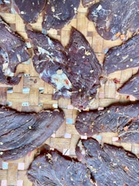 Slices of dried meat arranged on a woven bamboo mat, sprinkled with chili flakes and seasonings. The texture of the meat is rugged and fibrous.