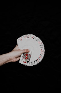 A hand is holding a fan of playing cards, spread out to reveal various numbers and suits. The focus is on a Jack of Spades, with red and black suits visible. The background is a dark black, creating a high contrast with the bright cards.