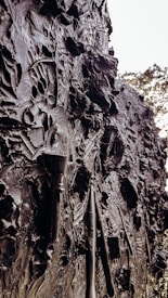 The image displays a textured wall embedded with various artistic tools. The surface is rough and uneven, with visible brush strokes and sculpting tools pressed into the material. The overall appearance is a dark, metallic or clay-like surface with intricate patterns and contours.