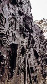 The image displays a textured wall embedded with various artistic tools. The surface is rough and uneven, with visible brush strokes and sculpting tools pressed into the material. The overall appearance is a dark, metallic or clay-like surface with intricate patterns and contours.