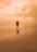 A solitary figure stands on a quiet beach with an expansive, cloudy sky overhead. The beach is wet, reflecting the person and the sky, creating a serene and surreal atmosphere.