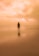 A solitary figure stands on a quiet beach with an expansive, cloudy sky overhead. The beach is wet, reflecting the person and the sky, creating a serene and surreal atmosphere.