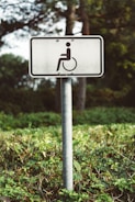 A white rectangular sign with a symbol of a person in a wheelchair is mounted on a metal pole. The sign is situated outdoors in a green vegetated area with trees and bushes in the background.