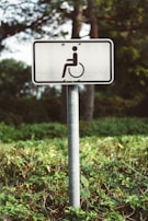 A white rectangular sign with a symbol of a person in a wheelchair is mounted on a metal pole. The sign is situated outdoors in a green vegetated area with trees and bushes in the background.