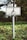 A white rectangular sign with a symbol of a person in a wheelchair is mounted on a metal pole. The sign is situated outdoors in a green vegetated area with trees and bushes in the background.