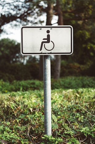 A white rectangular sign with a symbol of a person in a wheelchair is mounted on a metal pole. The sign is situated outdoors in a green vegetated area with trees and bushes in the background.