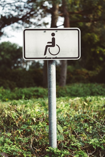 A white rectangular sign with a symbol of a person in a wheelchair is mounted on a metal pole. The sign is situated outdoors in a green vegetated area with trees and bushes in the background.