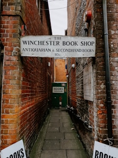 A narrow alleyway between two old brick buildings features a sign for 'The Winchester Book Shop,' which specializes in antiquarian and secondhand books. A green sign further down the alley directs toward books.