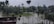 A landscape featuring a flooded area with partially submerged trees amid standing water. The sky is overcast, contributing to a gloomy atmosphere. A structure with a flat roof appears in the lower left corner, and tall utility poles or towers can be seen in the distance.