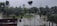 A landscape featuring a flooded area with partially submerged trees amid standing water. The sky is overcast, contributing to a gloomy atmosphere. A structure with a flat roof appears in the lower left corner, and tall utility poles or towers can be seen in the distance.