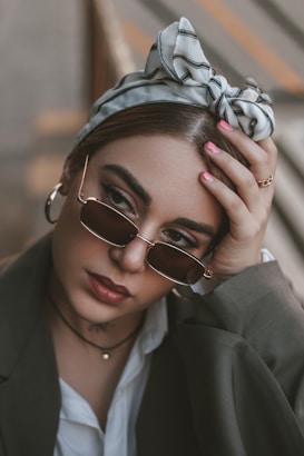 A person with stylish sunglasses and a patterned headscarf gazes thoughtfully, enhancing their fashion-forward appearance. They wear a dark blazer over a white shirt, accessorized with hoop earrings and a choker necklace. Their hand rests on their forehead, showcasing manicured nails.