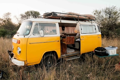 A vintage yellow camper van is parked in a grassy field, with the side door open revealing a cozy, lived-in interior. It has a roof rack loaded with camping equipment. The surroundings are natural, with tall dry grass and scattered bushes, creating a serene outdoor setting. A small table with cooking or camping gear is set up beside the van.