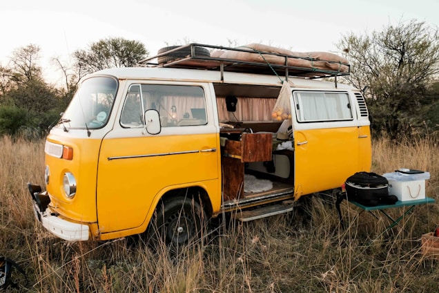 A vintage yellow camper van is parked in a grassy field, with the side door open revealing a cozy, lived-in interior. It has a roof rack loaded with camping equipment. The surroundings are natural, with tall dry grass and scattered bushes, creating a serene outdoor setting. A small table with cooking or camping gear is set up beside the van.