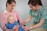 A nurse is administering a vaccine to a baby who is sitting on a woman's lap. The nurse is smiling and wearing colorful scrubs, while the woman gently holds the baby. The setting appears to be a medical office.