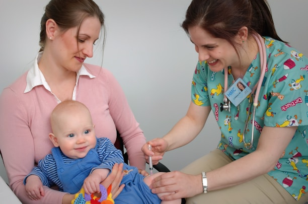 A nurse is administering a vaccine to a baby who is sitting on a woman's lap. The nurse is smiling and wearing colorful scrubs, while the woman gently holds the baby. The setting appears to be a medical office.