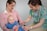 A nurse is administering a vaccine to a baby who is sitting on a woman's lap. The nurse is smiling and wearing colorful scrubs, while the woman gently holds the baby. The setting appears to be a medical office.
