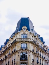 An ornate, historic building with a mansard roof, featuring detailed stonework and wrought iron balconies. The architecture exemplifies a classic, European style with multiple arched windows and elegant facades.