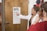 A woman in a white coat is pointing at an eye chart on a wooden door. Another person is facing her, possibly participating in an eye exam. The room contains a wooden bookshelf and some hats hanging on the wall.