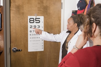 A woman in a white coat is pointing at an eye chart on a wooden door. Another person is facing her, possibly participating in an eye exam. The room contains a wooden bookshelf and some hats hanging on the wall.