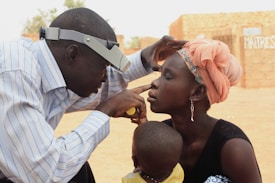 A man wearing a striped shirt and magnifying headset examines the eye of a woman who is sitting with her eyes closed. The woman is wearing a peach headscarf and a single long earring, holding a young child. The setting appears to be outdoors with brick walls in the background.