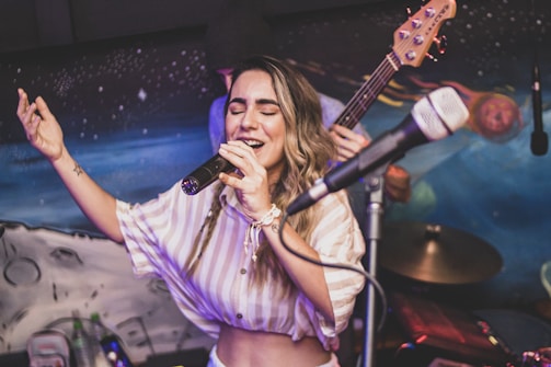 A woman is passionately singing into a microphone, with eyes closed, on a stage setting. She is wearing a striped top and has long wavy hair. In the background, there is musical equipment, including a bass guitar and a drum set. The backdrop is colorful and abstract, resembling a space or cosmic theme.