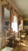 An ornate room featuring elaborately carved wooden furniture with gold accents, large mirrors, framed paintings, and sculptures. The room has an elegant, vintage flair with large windows adorned with intricate curtains that allow natural light to filter in. Chairs with white cushions are placed along a long, narrow carpet with intricate patterns.