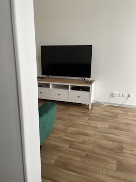 A modern living room setup with a large flat-screen television placed on a wooden TV stand with multiple drawers. The floor features light wooden flooring, and a partial view of a green sofa is visible. The wall is painted in a light color, and there are multiple electrical sockets nearby.