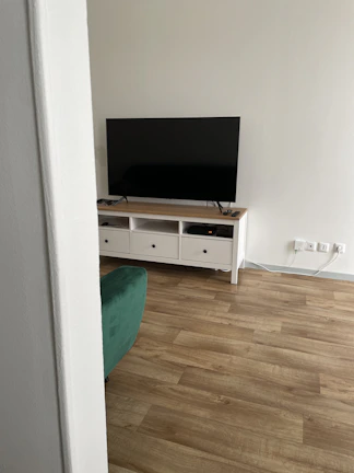 A modern living room setup with a large flat-screen television placed on a wooden TV stand with multiple drawers. The floor features light wooden flooring, and a partial view of a green sofa is visible. The wall is painted in a light color, and there are multiple electrical sockets nearby.