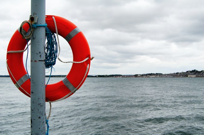 Red and white ring buoy with a coil of blue line hanging on a metal post at the water's edge, gray sea and a distant town under clouds
