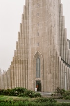 A tall, detailed concrete building with a series of staggered, vertical columns leading up to a narrow, pointed arch entrance. The architecture reflects a modern, geometric style. In front of the building, a small area of greenery with bushes and grass provides a contrasting natural element.