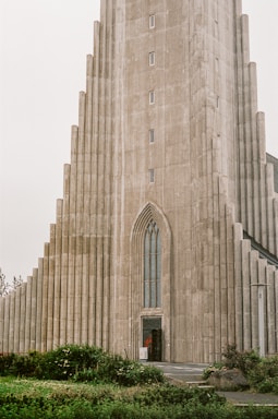A tall, detailed concrete building with a series of staggered, vertical columns leading up to a narrow, pointed arch entrance. The architecture reflects a modern, geometric style. In front of the building, a small area of greenery with bushes and grass provides a contrasting natural element.