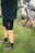A person wearing cycling attire, specifically black shorts and gray cycling shoes, is standing on grass next to a bicycle that is positioned against a white structure. The bicycle is equipped with bags, indicating it may be prepared for a long ride or cycling tour.