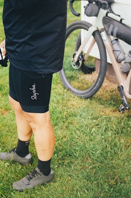 A person wearing cycling attire, specifically black shorts and gray cycling shoes, is standing on grass next to a bicycle that is positioned against a white structure. The bicycle is equipped with bags, indicating it may be prepared for a long ride or cycling tour.