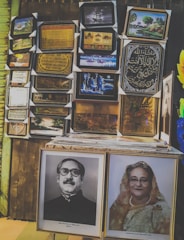 A variety of framed pictures are displayed on a wall, including images of landscapes, calligraphy, and portraits. A stack of frames sits on a table in front, with prominent black-and-white and colored portraits displayed.